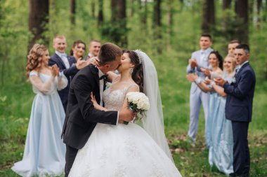 Bir grup insan izlerken bir gelin ve damat ormanda öpüşüyor. Gelin beyaz bir elbise giyiyor ve damat siyah bir takım giyiyor.