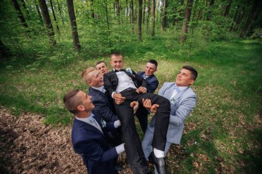 Bir grup adam ormanda oynuyor, biri diğerleri tarafından kaldırılıyor.