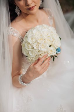 Bir gelin elinde bir buket beyaz gül tutuyor. Sahne büyük ihtimalle bir düğün ya da özel bir olay. Gelin beyaz giyinmiş.