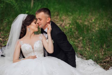 Bir gelin ve damat çimlerin üzerinde oturuyor, birbirlerini öpüyorlar. Gelin beyaz bir elbise giyiyor ve damat siyah bir takım elbise giyiyor. Sahne romantik ve samimi.