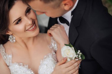 Bir gelin ve damat, damadın elinde beyaz bir gülle birbirlerini kucaklıyor. Gelin beyaz bir elbise giyiyor ve damat siyah bir takım elbise giyiyor. Sahne mutlu ve romantik.