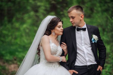 Gelin ve damat ormanda duruyorlar, gelin beyaz bir elbise ve damat siyah bir takım elbise giyiyor.