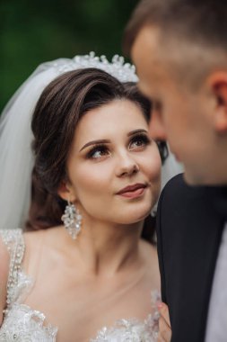 Bir gelin ve damat fotoğraf için poz veriyor. Damat siyah bir takım giyerken gelin beyaz bir elbise ve duvak takıyor. İkisi de gülümsüyor ve kameraya bakıyor.