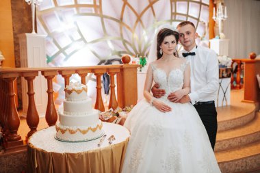 Bir gelin ve damat düğün pastasının önünde durur. Gelin beyaz bir elbise giyiyor ve damat siyah bir takım elbise giyiyor. Pasta altın ve beyaz kremayla süslenmiş.