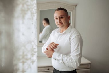 Beyaz gömlekli bir adam aynanın önünde duruyor, gömleğini düzeltiyor. Özgüven kavramı ve detaylara dikkat etme, adam görünüşünün temiz ve düzenli olduğundan emin olmak için zaman ayırıyor.