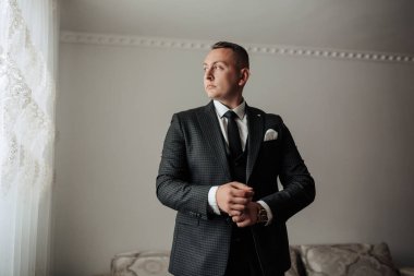 Takım elbiseli bir adam pencerenin önünde duruyor, kravatını düzeltiyor. Adam takım elbise ve kravat giydiği için resmiyet ve profesyonellik kavramı. Pencere dış dünyayı gösteriyor.