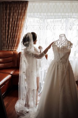 Bir kadın evlenmeye hazırlanıyor ve gelinliğine duvak takıyor. Elbise beyazdır ve bir pencerede sergilenir