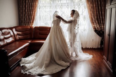 Bir gelin düğünü için hazırlanıyor ve bir elbisenin önünde duruyor. Bir kadın hazırlanmasına yardım ediyor.