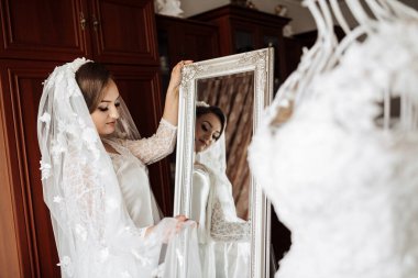 Bir kadın düğünü için hazırlanıyor ve aynada kendine bakıyor. Ayna beyaz bir elbisenin yanında.