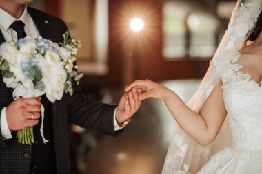 Bir gelin ve damat el ele tutuşuyor ve gelin bir buket tutuyor. Gelin beyaz bir elbise giyiyor ve damat bir takım elbise giyiyor. Sahne muhtemelen bir düğün merasimi.