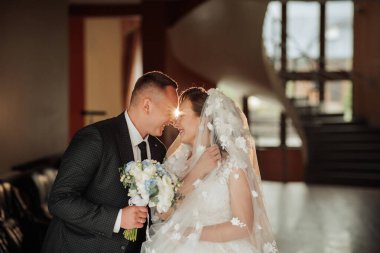 Bir gelin ve damat kilisede sarılıyor. Gelin beyaz bir elbise ve duvak takıyor. Gelin elinde bir buket çiçek tutuyor.