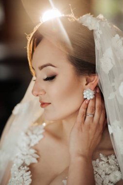 Beyaz bir duvak ve küpe takan bir kadın. Kameraya bakıyor.