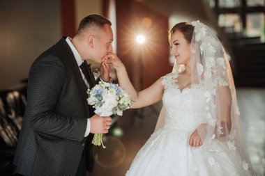 Bir gelin ve damat birbirlerinin alnını öpüyor. Gelin elinde bir buket çiçek tutuyor.