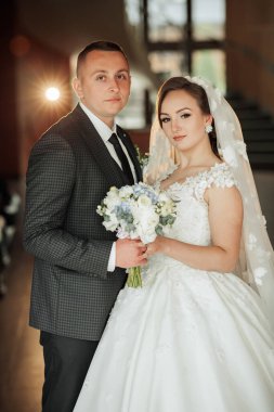 Bir gelin ve damat fotoğraf için poz veriyor. Gelin beyaz bir elbise giyiyor ve elinde bir buket çiçek tutuyor. Aşk ve mutluluk kavramı