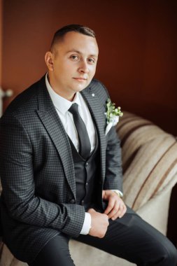 Takım elbiseli ve kravatlı bir adam kanepede oturur. Kravat ve yelek giyiyor.