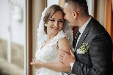 Bir gelin ve damat pencerede birbirlerine sarılıyorlar. Gelin beyaz bir elbise giyiyor ve damat bir takım elbise giyiyor. Sahne mutlu ve sevgi dolu.