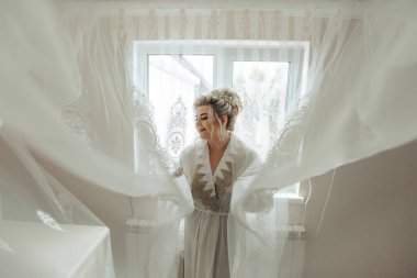 Beyaz elbiseli bir kadın pencerenin önünde duruyor, beyaz bir perdeyle örtülmüş. Özel bir durum için hazırlanıyor, muhtemelen bir düğün için. Zarafet ve beklenti kavramı