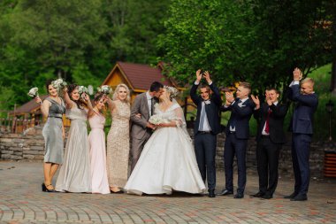 Bir grup insan bir resim için poz veriyor, bir gelin ve damat da dahil. Gelin beyaz bir elbise giyiyor ve damat bir takım elbise giyiyor. Grup bir binanın önünde duruyor.