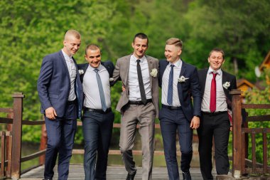 Takım elbiseli ve kravatlı beş adamdan oluşan bir grup köprüde fotoğraf için poz veriyor. Sahne neşeli ve eğlenceli, çünkü erkekler gülümsüyor ve birbirlerinin arkadaşlığından zevk alıyorlar.