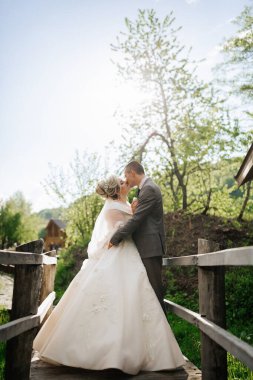Bir gelin ve damat köprüde öpüşüyor. Gelin beyaz bir elbise giyiyor ve damat bir takım elbise giyiyor. Sahne romantik ve samimi.