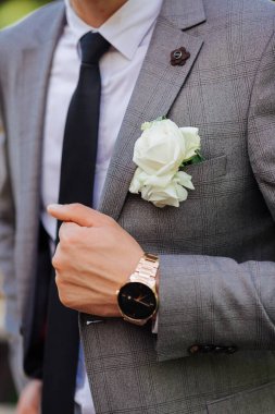 Takım elbiseli ve kravatlı bir adam saat takıyor ve elinde beyaz bir gül tutuyor.