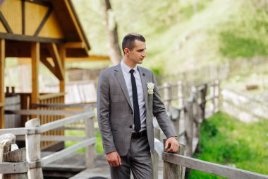 Takım elbiseli ve kravatlı bir adam ahşap bir köprüde duruyor. Elinde beyaz bir çiçek tutuyor.