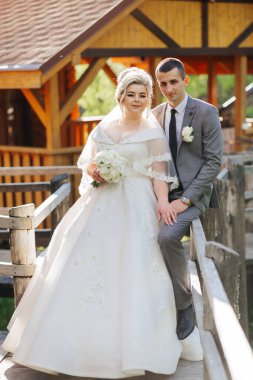 Gelin ve damat ahşap bir köprüde fotoğraf için poz veriyorlar. Gelin beyaz bir elbise giyiyor ve damat takım elbise ve kravat giyerken elinde bir buket tutuyor. Sahne mutlu ve romantik.