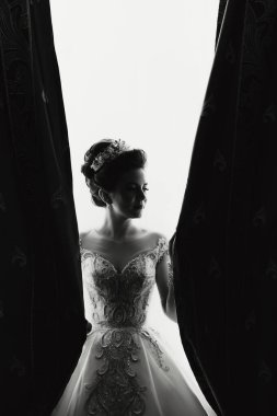 Gelinlikli bir kadın perdenin önünde duruyor. O bir taç takıyor ve saçları topuz şeklinde. İmajın romantik ve zarif bir havası var.