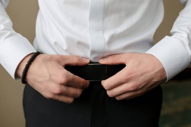 Bir adam beyaz gömlek giyerken kemerini ayarlıyor. Formalite ve detaylara dikkat etme kavramı. Çünkü adam kemerinin düzgün bağlandığından emin olmak istiyor.