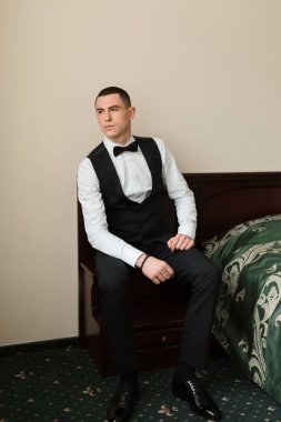 Siyah yelek ve beyaz gömlekli bir adam bir yatağa oturur. Siyah papyon ve siyah kuşak takıyor.