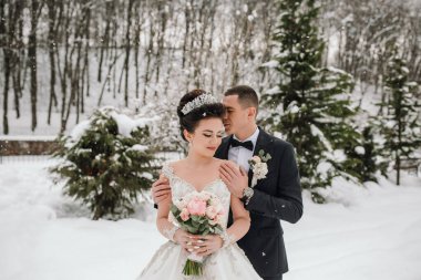 Bir gelin ve damat karda fotoğraf için poz veriyorlar. Damat takım elbise giyerken gelin beyaz bir elbise ve taç giyiyor. Etrafları ağaçlar ve karlarla çevrili. Romantik bir ortam yaratıyorlar.