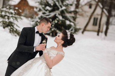 Bir gelin ve damat karda fotoğraf için poz veriyorlar. Gelin beyaz bir elbise giyiyor ve damat siyah bir takım elbise giyiyor. İkisi de gülümsüyor ve kameraya bakıyor.