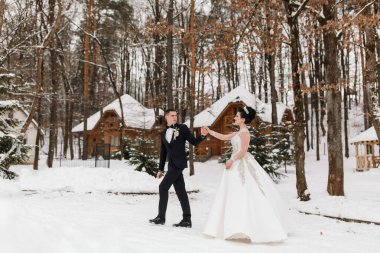 Bir gelin ve damat bir çadırın önünde karda yürüyorlar. Gelin beyaz bir elbise giyiyor ve damat bir takım elbise giyiyor. Sahne romantik ve resimli.