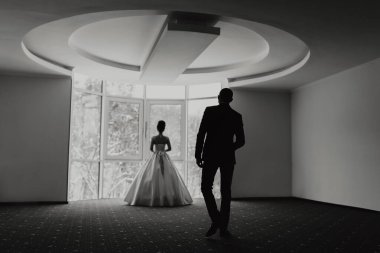 Bir adam ve bir kadın pencereli bir odada duruyorlar. Adam takım elbise giyiyor ve kadın da elbise giyiyor. Sahne romantik ve samimi.