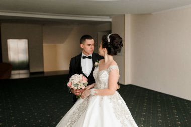 Gelin ve damat büyük bir odada duruyorlar, gelin beyaz bir elbise giyiyor ve damat siyah bir takım elbise giyiyor.