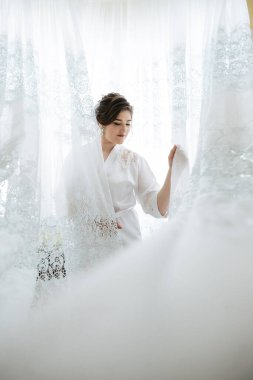 Beyaz cüppeli bir kadın büyük beyaz bir elbisenin önünde duruyor. Elbise onun üzerine örtülmüş ve o elbiseye uzanıyor. Zarafet ve incelik kavramı