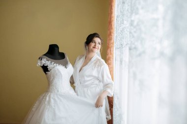 Bir kadın gelinliğin önünde duruyor, yüzüne dayayıp duruyor. Elbise beyaz ve dantelli. Kadın elbiseye hayran.