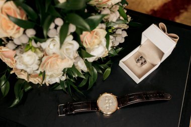 Masada bir buket çiçeğin ve beyaz bir kutunun yanında bir saat var. Bu saat altın ve siyah.