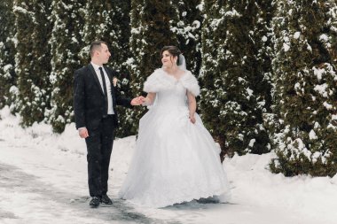 Gelin ve damat karda yürüyorlar, gelin beyaz bir elbise ve damat siyah bir takım elbise giyiyor.