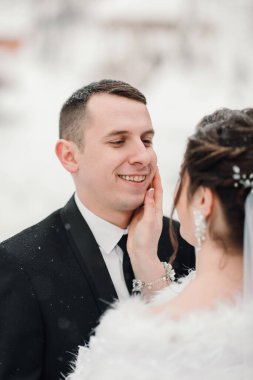 Bir adam ve bir kadın kameraya gülümsüyor. Adam takım elbise ve kravat takıyor ve kadın beyaz bir kürk ceket giyiyor. İkisi de kameraya bakıyor ve mutlu görünüyorlar.