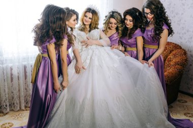 Bir grup kadın gelinlik giymiş bir gelinin etrafında toplanıyor. Gelin beyaz bir elbise giyiyor ve nedimeleriyle çevrili. Kadınların hepsi mor giyinmiş.