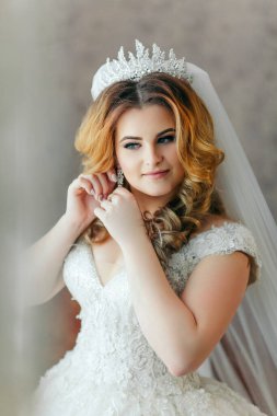 Gelinlikli bir kadın saçını yaptırıyor. Bir taç ve küpe takıyor.