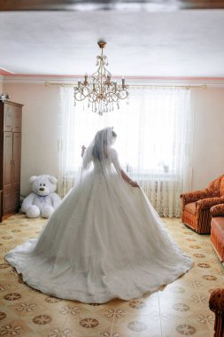 Beyaz elbiseli bir gelin oyuncak ayının önünde duruyor. Oda bir avize ve kanepe ile süslenmiş.