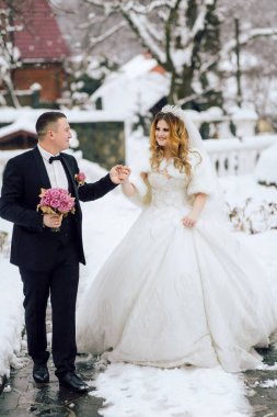 Bir gelin ve damat karda yürüyor, gelin kürk şort giyiyor. Gelin elinde bir buket çiçek tutuyor.