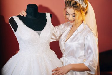 Bir gelin gelinliğin yanında duruyor, gülümsüyor. Elbise beyaz ve bir mankenin üzerine sarılı.