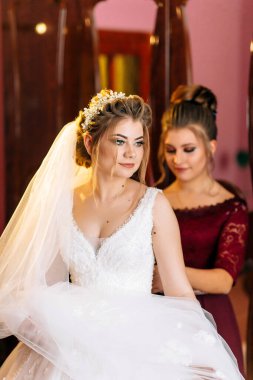 Bir gelin, nedimesi ona yardım ederken düğününe hazırlanıyor. Gelin beyaz bir elbise ve taç giyiyor ve nedime de kırmızı bir elbise giyiyor.