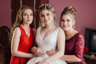 Üç kadın fotoğraf için poz veriyor, biri gelinlik giyiyor. Diğer iki kadın kırmızı elbise giyiyor. Sahne mutlu ve kutlama dolu.