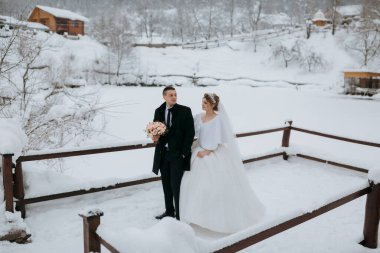 Bir gelin ve damat, damat elinde buketle, karlı bir yolda dikiliyorlar. Sahne, arka planında ahşap bir çitle bir kış harikalar diyarında geçiyor.