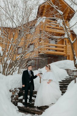 Bir gelin ve damat, ahşap bir evin önündeki karla kaplı merdivenlerden iniyorlar. Gelin beyaz bir elbise giyiyor ve damat siyah bir takım elbise giyiyor. Sahne romantik ve samimi.