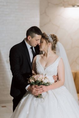 Bir gelin ve damat bir buket çiçek tutarken birbirlerine sarılıyorlar. Gelin beyaz bir elbise giyiyor ve damat bir takım elbise giyiyor. Sahne mutlu ve romantik.